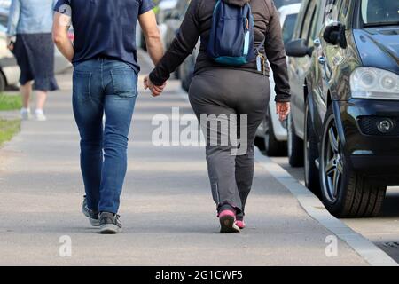 Fat woman in sportswear and slim man in jeans walking along a city street with parked cars. Couple in love holding hands, summer fashion