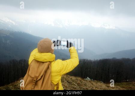Tourist in yellow jacket with backpack doing selfie in spring snowy mountains. Travel concept. Landscape photography Stock Photo