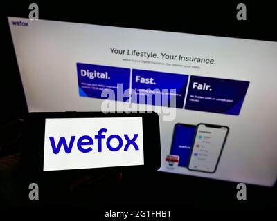 Person holding smartphone with logo of digital insurance company wefox Holding AG on screen in front of website. Focus on phone display. Stock Photo