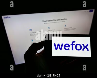 Person holding mobile phone with logo of digital insurance company wefox Holding AG on screen in front of business web page. Focus on phone display. Stock Photo