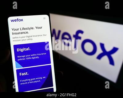 Person holding mobile phone with website of digital insurance company wefox Holding AG on screen in front of logo. Focus on center of phone display. Stock Photo