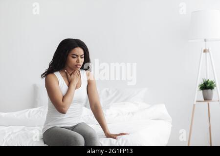 Pharyngitis. Sick black woman touching sore throat, feeling unwell and coughing, sitting on bed at home, free space Stock Photo