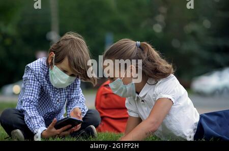 Children sitting on grass with mobile devices. Cute pupils using smartphones at the elementary school. Boy and girl in safety masks with gadgets in their hands. Dependence on the mobile phone. Stock Photo