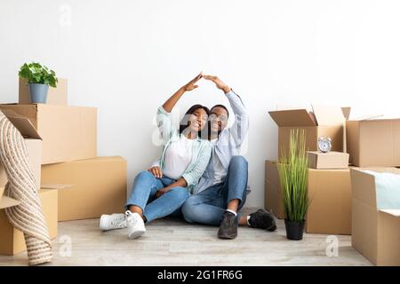 Mortgage for young families. Happy black couple moving to their own home, joining hands making symbolic house roof Stock Photo