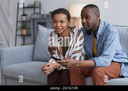 Portrait of modern African-American couple watching TV or movies while sitting on sofa at home, copy space Stock Photo