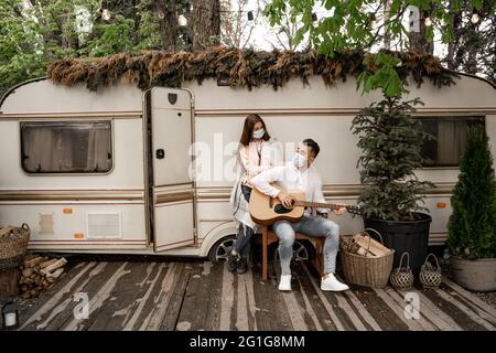 man in medical mask playing guitar near girlfriend and trailer in camping Stock Photo