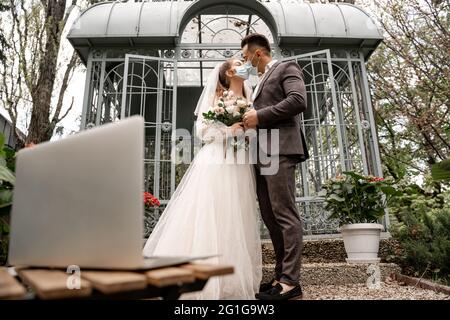 young newlyweds in medical masks kissing outdoors near laptop on blurred foreground Stock Photo