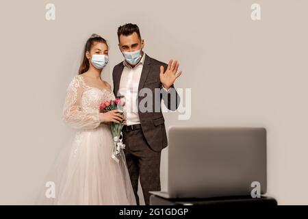 groom in medical mask waving hand during video call on blurred laptop isolated on grey Stock Photo