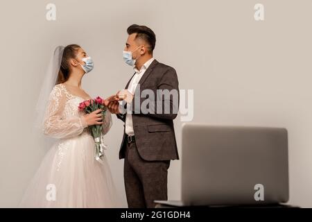 young newlyweds in medical masks looking at each other near blurred laptop isolated on grey Stock Photo