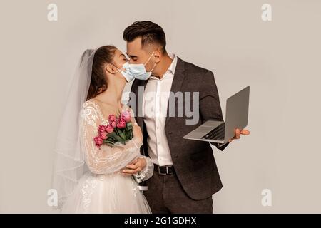 man in medical mask kissing bride while holding laptop isolated on grey Stock Photo
