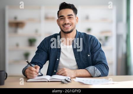 Cheerful arab guy in casual sitting at workdesk, taking notes Stock Photo