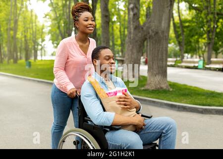 Young black disabled man in wheelchair holding paper bag with products, going shopping with his girlfriend Stock Photo