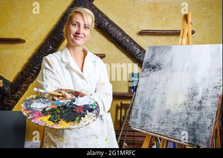 Satisfied female artist posing near new picture next to easel. High quality photo. Stock Photo