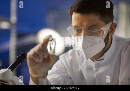 Portrait of male scientist in goggles and face mask looking at glass vial in hand Stock Photo