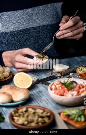 a young caucasian man, sitting at a gray rustic wooden table, puts some mushrooms fried with garlic and parsley on a halved bread bun Stock Photo