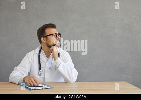 Dreamful pensive male doctor in white medical gown suit sit at desk look aside Stock Photo