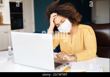 Suffering from headache African-American woman in protective medical mask, tired and weary female employee sits in front of laptop with eyes closed, holds head, feels viral disease symptoms Stock Photo