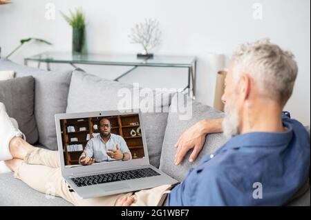 Back view over shoulder of mature man at the laptop screen with African-American man on it, two diverse male colleagues talking online, using computer app for video connection Stock Photo
