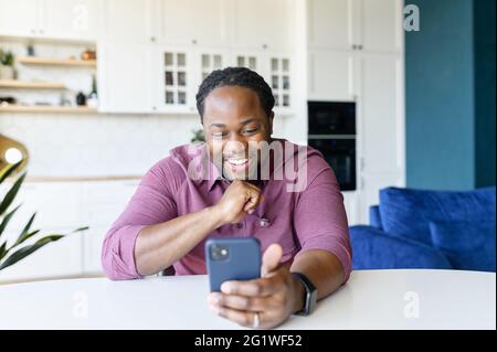 Overjoyed African-American guy enjoys spending time online in social networks, sitting at the desk at home and using smartphone, messaging and chatting, black man holds mobile and laughs Stock Photo