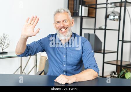 Cheerful middle-aged male employee in smart casual shirt looks at the camera and waving hi, greeting participants of online meeting. Video chat with happy senior man Stock Photo