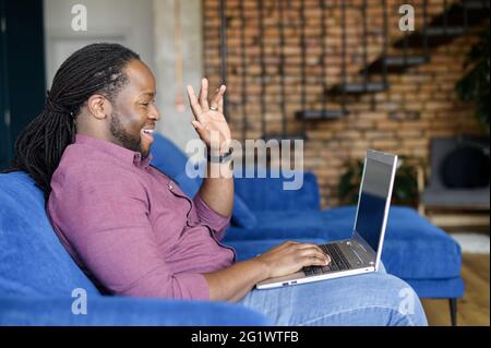 Smiling and happy African-American guy makes video call on the laptop, waving hello, greeting family or colleagues on the distance, using new computer app for video connection, takes part in webinar Stock Photo