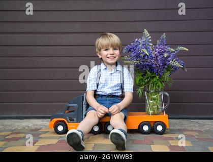 funny cute 4-5 year old boy in shorts and a shirt sits on a toy orange truck with a large bouquet of lupins flowers. little courier, a gift for women, Stock Photo