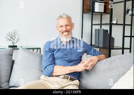 Handsome optimistic mature man staring at the camera with cheerful toothy smile, happy senior guy in smart casual wear sitting on the sofa at home. Lifestyle and wellbeing concept Stock Photo