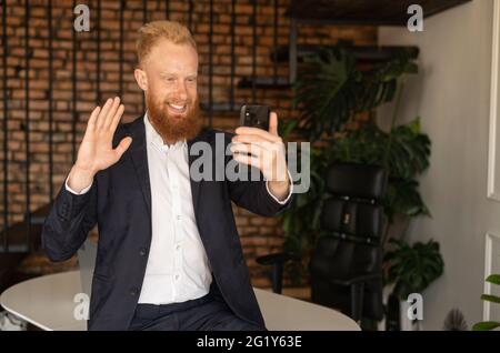Cheerful contemporary young red-haired businessman in formal suit using smartphone for making video call, waving, greeting colleagues or partners, male employee using mobile app for video connection Stock Photo