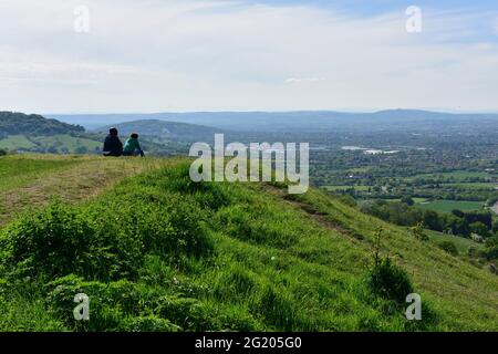 Two people sitting on grassy escarpment above Severn Vale looking across Gloucester toward Malvern Hills, Gloucestershire, UK Stock Photo