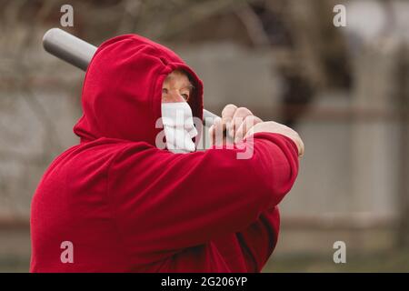 Angry aggressive elderly man in protective safe medical mask swings baseball bat in the background of outdoor street, portrait, close up. Stock Photo