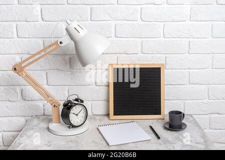 Table lamp, empty blank letter board, notebook and alarm clock on the table, against the background of a white brick wall. Workplace, front view, mock Stock Photo