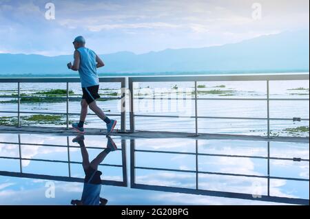 Motion blur. An Asian senior man in fitness wear running in a park along a lake at sunrise, blue mountains blurred in the background. Stock Photo