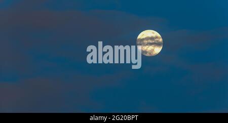 Full moon hiding behind the ripples clouds on the blue sky at dusk. Stock Photo