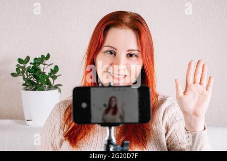 Young red-haired woman blogger leads an online stream using a smartphone. Influencer greets followers. The modern businesswoman works from home using Stock Photo