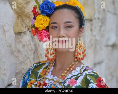 Young beautiful Mexican woman with makeup wears traditional Mayan Yucatecan folkloric dress with flowers in her hair and poses for the camera. Stock Photo