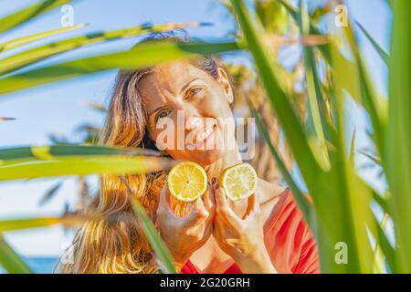 Woman portrait holding two lemon slices between palm tree leaves.Colorful healthy fruit concept background. Stock Photo