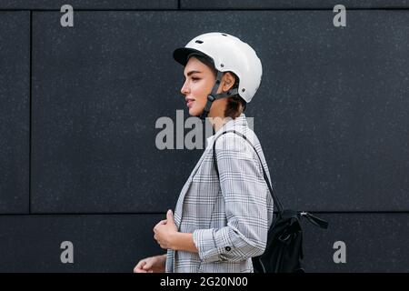 Side view of a good looking businesswoman in cycling helmet standing at black wall in the city Stock Photo