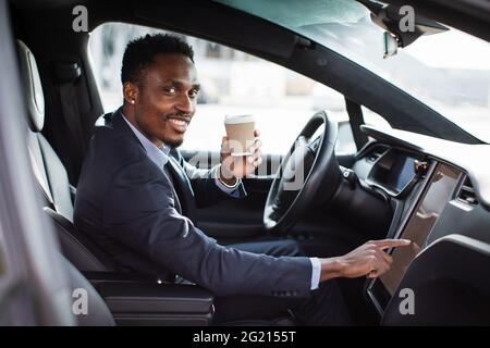 Smiling businessman in suit drinking coffee and using gps navigator at his modern luxury car. Happy african male tapping with finger on dashboard of his electric car. Stock Photo