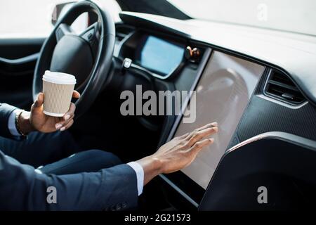Close up african man in suit sitting on driver's seat of electric car and touching dashboard with finger. Young man using modern system of luxury vehicle for navigation. Stock Photo