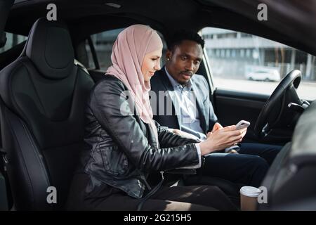 African american businessman in suit and muslim woman in hijab using modern smartphone for remote work while sitting together inside luxury car. Concept of people, gadgets and vehicle. Stock Photo