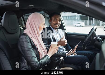 African american businessman in suit and muslim woman in hijab using modern smartphone for remote work while sitting together inside luxury car. Concept of people, gadgets and vehicle. Stock Photo