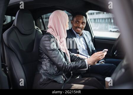 African american businessman in suit and muslim woman in hijab using modern smartphone for remote work while sitting together inside luxury car. Concept of people, gadgets and vehicle. Stock Photo