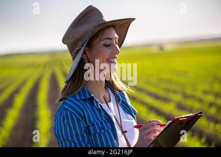 Attractive young farmer smiling standing in corn field in spring. Female agronomist taking notes using clipboard. Stock Photo