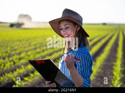 Portrait of female agronomist taking notes on growth of corn. Young farmer standing in field smiling satisfied. Stock Photo