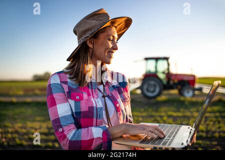 Female agronomist using laptop modern technology in agriculture. Young farmer standing in field in front of tractor machinery smiling. Stock Photo
