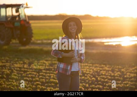 Young woman farmer looking at camera in field holding clipboard. Agronomist standing on plantation in front of tractor machinery. Stock Photo