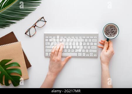 Woman typing on computer keyboard and drinking coffee at workplace. Flat lay office desktop workspace with female hands on white background Stock Photo