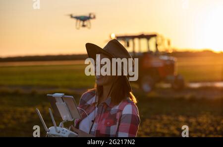 Woman agronomist using drone modern technology in agriculture. Young farmer standing in field at sunset in front of tractor machinery. Stock Photo