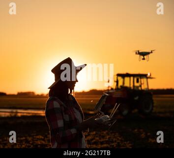 Female agronomist flying drone using controller innovation in agriculture. Young farmer standing in field with tractor in background at sunset. Stock Photo