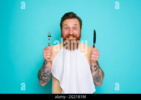 Happy man with tattoos is ready to eat with cutlery in hand Stock Photo